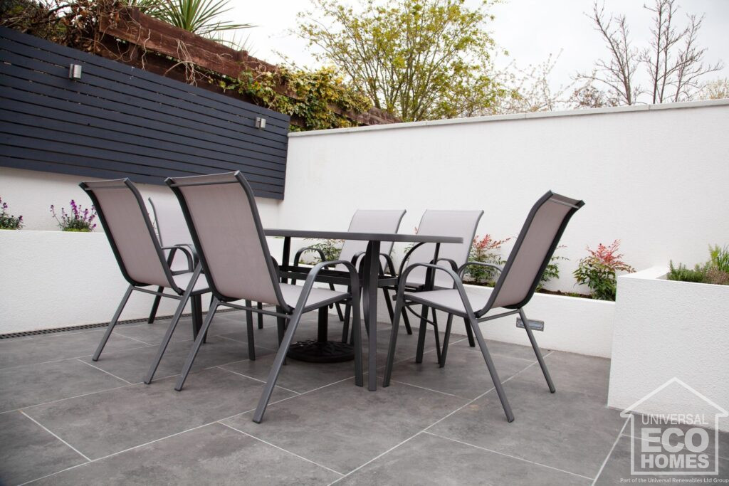 A modern outdoor patio with a dining table set and surrounding plant beds under an overcast sky.